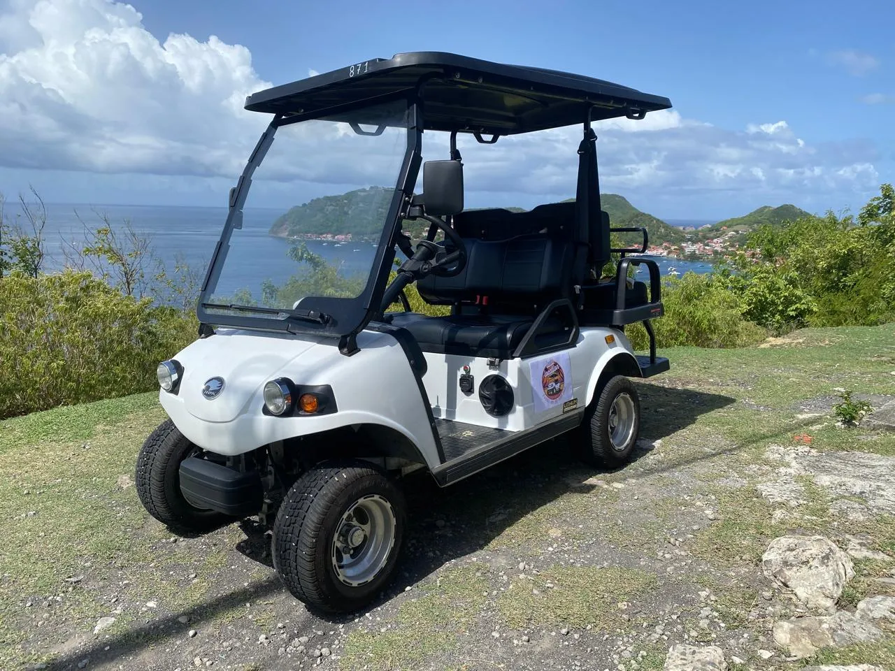 Voiturette blanche avec vue sur la mer et collines verdoyantes.
