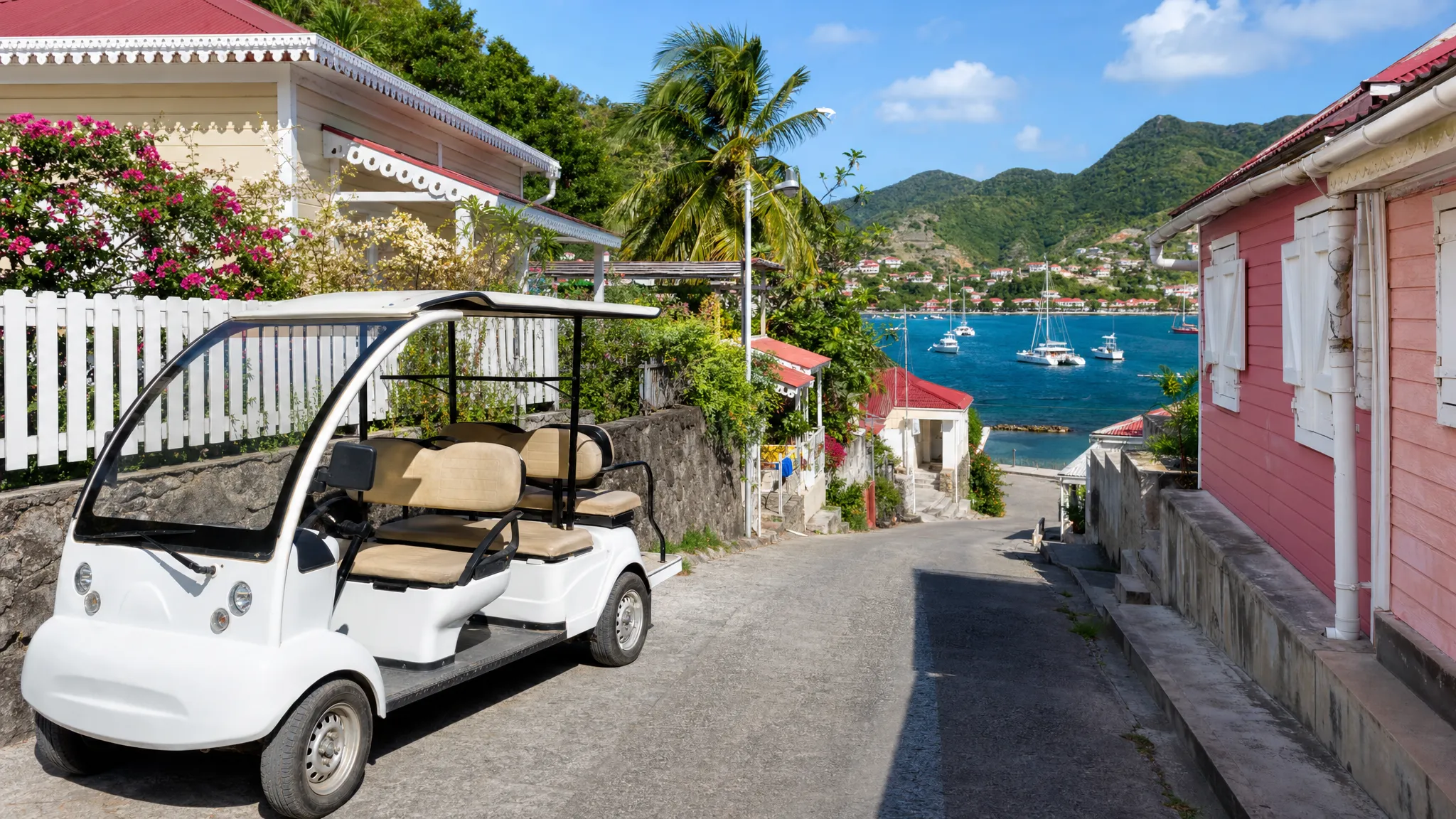 Une voiturette &eacute;lectrique blanche stationn&eacute;e pr&egrave;s d&rsquo;une ruelle tranquille &agrave; Terre-de-Haut, avec des maisons cr&eacute;oles color&eacute;es, des palmiers et la baie des Saintes en arri&egrave;re-plan.