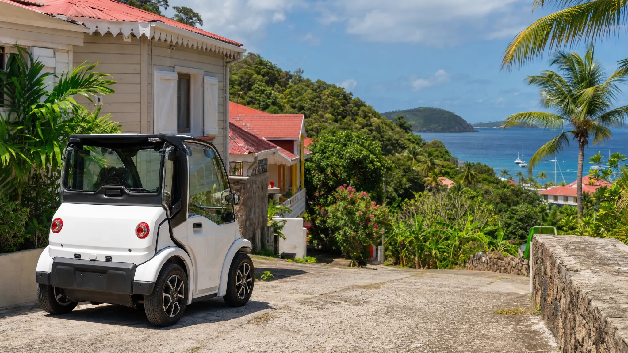Une voiturette &eacute;lectrique compacte stationn&eacute;e pr&egrave;s d&rsquo;un chemin tranquille &agrave; Terre-de-Haut, avec quelques maisons cr&eacute;oles, de la v&eacute;g&eacute;tation tropicale et la mer au loin.