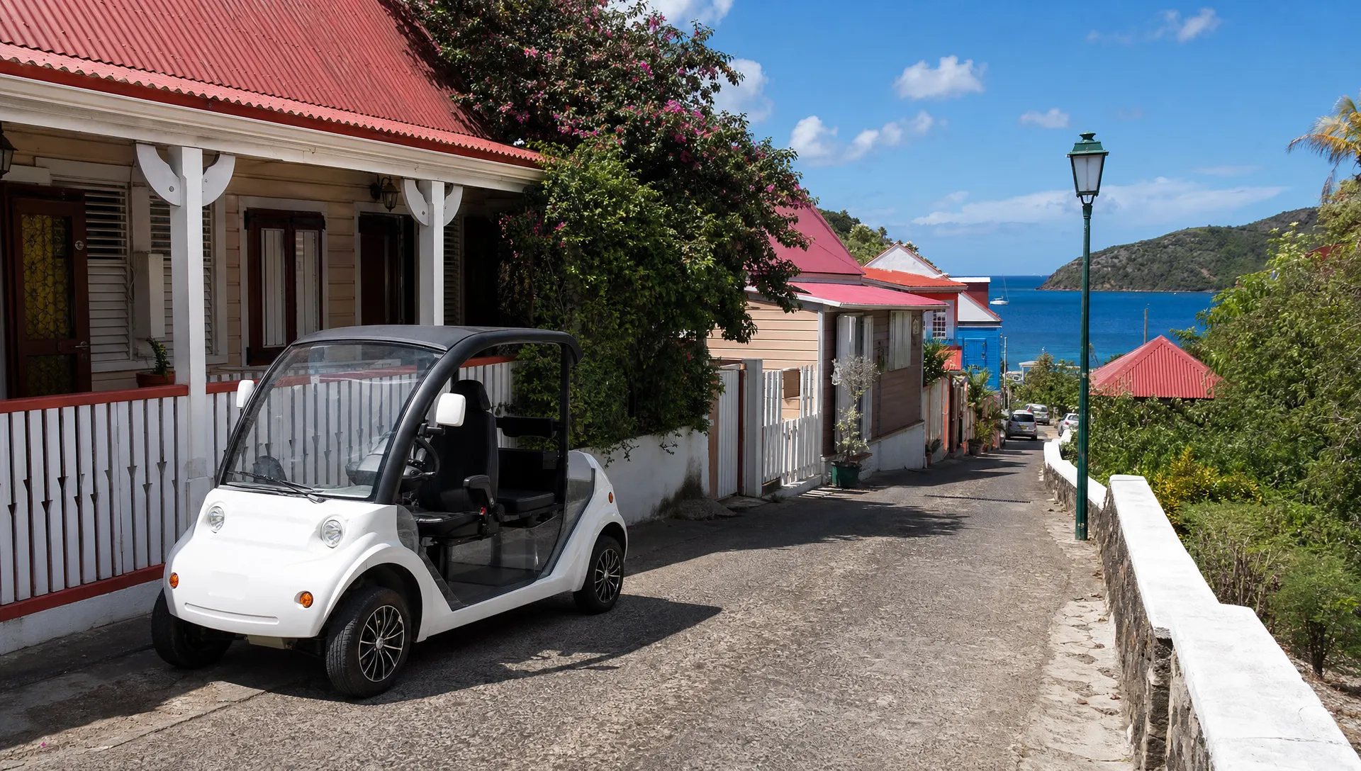 Une voiturette électrique blanche garée le long d’une rue étroite de Terre-de-Haut, avec des maisons créoles colorées, un muret en bord de route et la mer visible au loin.