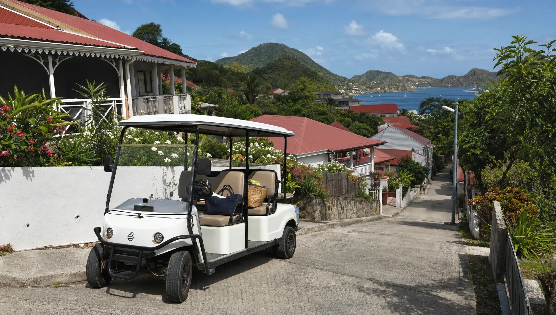 Une voiturette &eacute;lectrique blanche gar&eacute;e sur une route en pente &agrave; Terre-de-Haut, avec des maisons cr&eacute;oles, des collines vertes et la mer des Saintes au loin, quelques sacs de plage visibles &agrave; bord.