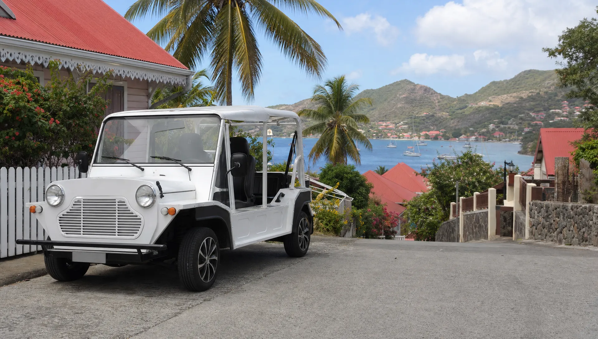 Une voiturette électrique blanche est garée dans une rue calme de Terre-de-Haut, avec des maisons créoles, des palmiers et la baie des Saintes derrière elle.