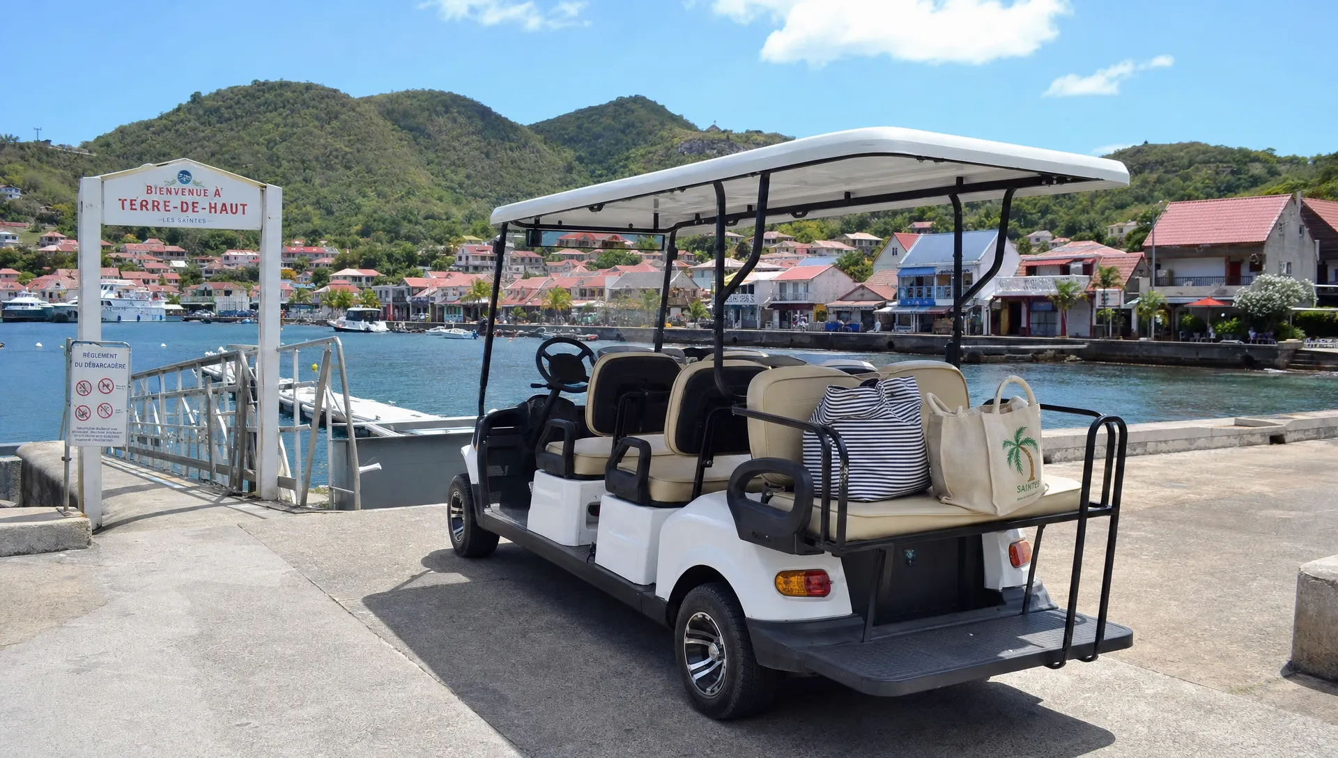 Une voiturette &eacute;lectrique blanche pr&egrave;s du d&eacute;barcad&egrave;re de Terre-de-Haut, avec des sacs l&eacute;gers &agrave; bord et le bourg des Saintes derri&egrave;re.