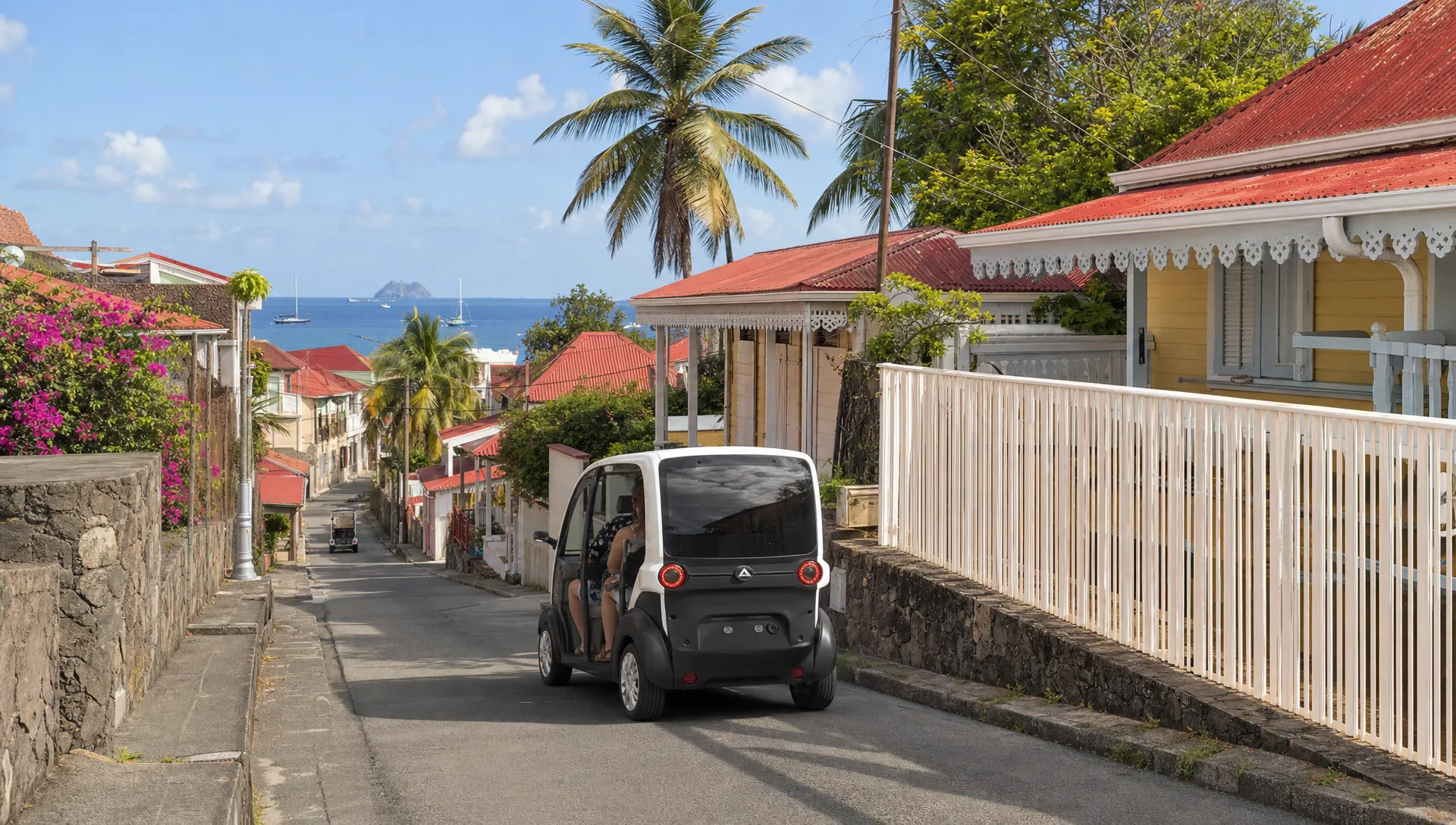 Une voiturette &eacute;lectrique compacte circule dans une rue de Terre-de-Haut, avec des maisons cr&eacute;oles, des palmiers et la mer au loin.