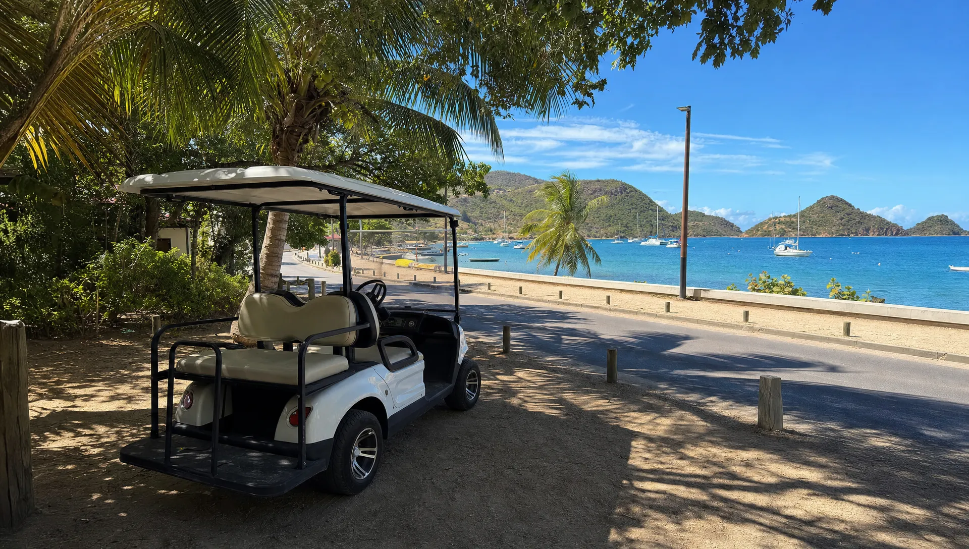 Une voiturette &eacute;lectrique stationn&eacute;e &agrave; l&rsquo;ombre pr&egrave;s d&rsquo;une plage de Terre-de-Haut, avec des palmiers, une route calme et la mer des Saintes au fond.